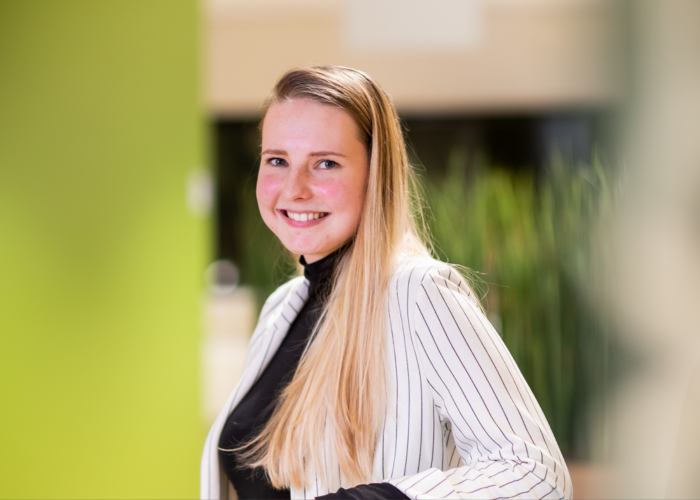 Student Anne Meijboom van de Associate degree Human Resource Management draagt een wit colbert met krijtstreep en poseert bij een limegroene muur.