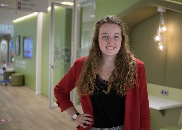 Student Lianne Groenendijk draagt een rode blazer maar daaronder een zwart shirt. Ze staat in het schoolgebouw voor een groene achtergrond.